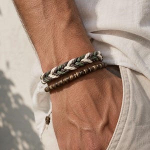 May include: A set of two bracelets consisting of one braided cord bracelet in shades of white, dark green, and brown, and one bracelet featuring small, cylindrical, dark brown wooden beads arranged in a single continuous row.