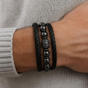May include: A set of three black bracelets. Two are braided leather, and the center one features a Buddha head and dark beads. The bracelets are worn on a person's wrist, with a gray sweater in the background.