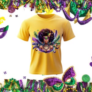 以下が含まれることがあります： マスタードイエローのTシャツ。仮面舞踏会のマスクをつけた女性の鮮やかなグラフィックが特徴で、羽と宝石で飾られています。デザインには紫、緑、金色の要素が組み込まれており、お祝いのマルディグラのテーマを連想させます。装飾的なビーズとマスクがシャツを囲んでいます。