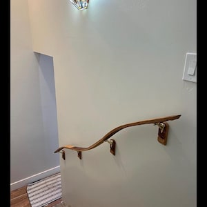 May include: A wooden handrail with a curved design, mounted on a light-colored wall. The handrail is supported by decorative brackets. A stained glass light fixture is mounted on the wall above the handrail.