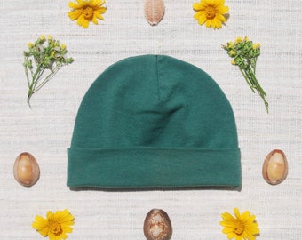 May include: A dark green beanie hat on a white textured background with yellow flowers and brown seed pods.