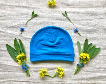 May include: A bright blue beanie hat on a white linen background with yellow and blue flowers.