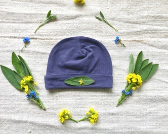 May include: A dark blue knitted baby hat with a green leaf detail. The hat is surrounded by small yellow and blue flowers on a white fabric background.