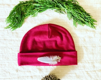 May include: A crimson red beanie with a white feather accent. The beanie is made of a soft, comfortable fabric and has a simple, classic design.
