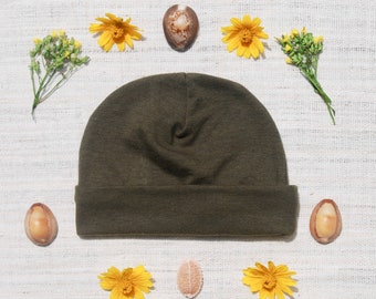May include: A dark green beanie hat with a folded brim, surrounded by yellow flowers and seashells on a white textured background.