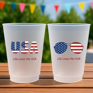 May include: Two frosted plastic cups with patriotic designs. One cup features the word "USA" in red, white, and blue. The other cup displays sunglasses with a star-spangled lens and striped lens. Both cups have the text "Lila Loves Her USA".