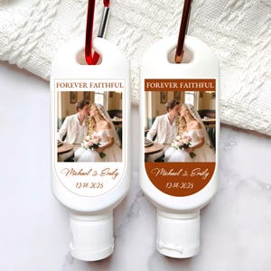 May include: Two white hand sanitizer bottles with red carabiner clips. Each bottle has a label with a wedding photo, the text "FOREVER FAITHFUL", and the names "Michael & Emily" with the date "12.14.2025".
