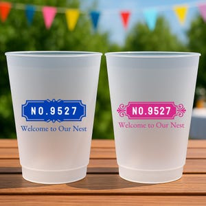 May include: Two frosted plastic cups with the text "NO.9527 Welcome to Our Nest" in blue and pink. The cups are on a wooden table with colorful flags in the background.