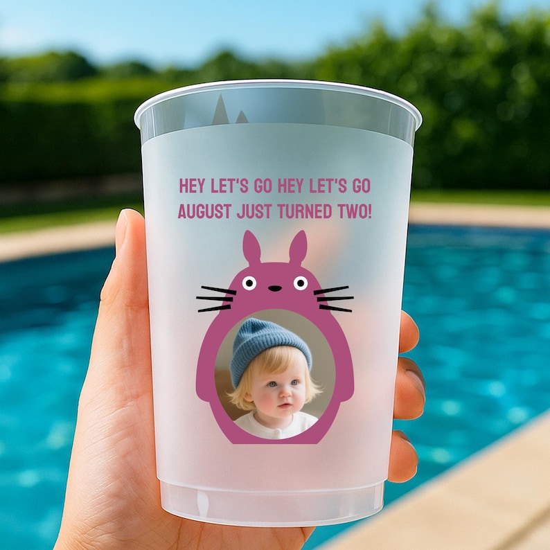 May include: A clear plastic cup with a frosted finish. The cup features a purple cartoon character with a photo of a baby inside. Text on the cup reads "HEY LET'S GO HEY LET'S GO AUGUST JUST TURNED TWO!"