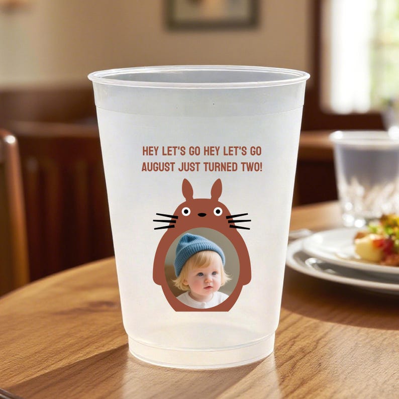 May include: A clear plastic cup with the text "HEY LET'S GO HEY LET'S GO AUGUST JUST TURNED TWO!" and a cartoon illustration of a character holding a photo of a child wearing a blue beanie. The cup is sitting on a wooden table.