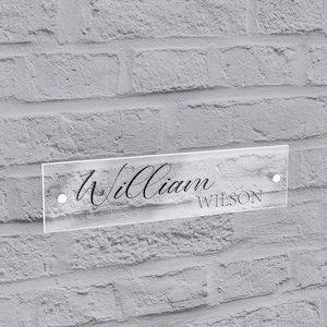 May include: A clear acrylic sign with the name "William" in elegant script above the last name "Wilson" in a smaller, sans-serif font. The sign is mounted on a gray brick wall with two silver-colored screws.