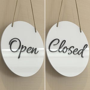 May include: A white, circular sign with the word "Open" in black script, hanging by a rope. The sign is displayed next to a similar sign with the word "Closed". The background is a light wood grain.