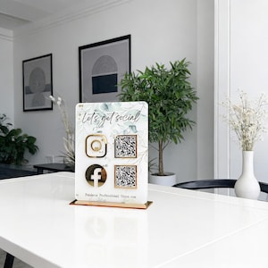 Puede incluir: Un letrero de redes sociales de acrílico transparente con detalles dorados. El letrero muestra el texto "Let's get social" e iconos de Instagram y Facebook, además de códigos QR. La base es de color marrón claro. El letrero está sobre una mesa blanca.
