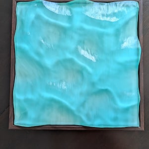 May include: A square wall art piece featuring a translucent, wavy turquoise glass panel set within a dark brown wooden frame. The glass surface has a rippled, undulating texture that creates light reflections, mimicking the appearance of water.