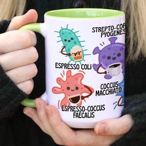 May include: White ceramic mug with a green interior and handle, featuring cartoon bacteria illustrations holding coffee cups. Text includes "Espresso Coli", "Strepto-Coc Pyogenes", and "Espresso-Coccus Faecalis".