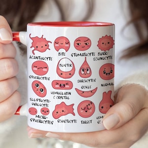 Op de afbeelding: Witte keramische mok met een rode binnenkant en handvat, met cartoon illustraties van bloedcellen met verschillende uitdrukkingen en gelabeld met wetenschappelijke termen zoals "Acanthocyte" en "Spherocytes".