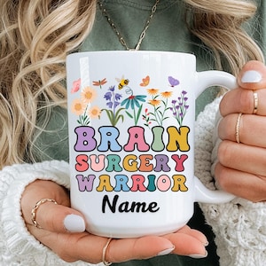 May include: White ceramic mug with the text "BRAIN SURGERY WARRIOR Name" in colorful, bubble letters. The mug is decorated with a floral design featuring butterflies, bees, and various wildflowers. The mug has a white handle.