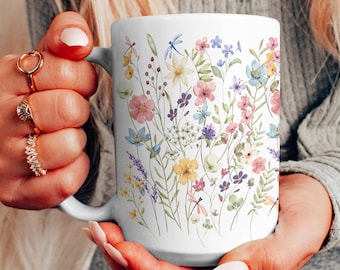 Può includere: Tazza in ceramica bianca con un colorato motivo floreale con vari fiori selvatici e una libellula. La tazza ha un manico curvo. Il motivo floreale comprende fiori rosa, blu, gialli e viola.