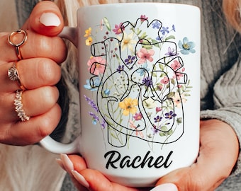 May include: White ceramic mug featuring a floral heart design with colorful flowers. The mug has the name "Rachel" printed in black script. A unique and artistic gift idea.