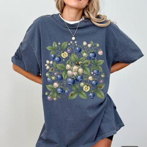 May include: A blue-grey t-shirt with a botanical illustration of blueberries, leaves, and flowers. The design includes a bee and ladybirds. The shirt has short sleeves and a loose fit. The word "DEMIN" is printed on the bottom right.