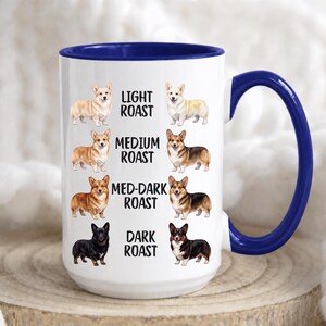 May include: A white ceramic mug with a blue interior and handle, featuring illustrations of Corgi dogs arranged in rows next to the text Light Roast, Medium Roast, Med-Dark Roast, and Dark Roast, depicting various coat colors and patterns.