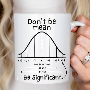 Könnte beinhalten: Weiße Keramik-Tasse mit dem Text "Don't be mean, Be Significant" und einem Glockenkurvendiagramm. Die Tasse wird von einer Person gehalten.
