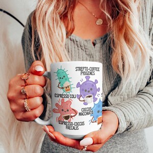 May include: A white ceramic coffee mug featuring cartoon illustrations of bacteria, each holding a coffee cup. The mug includes the text "Strepto-Coffee Pyogenes," "Espresso Coli," "Cocus Macchiato," and "Espresso-Cocus Faecalis."