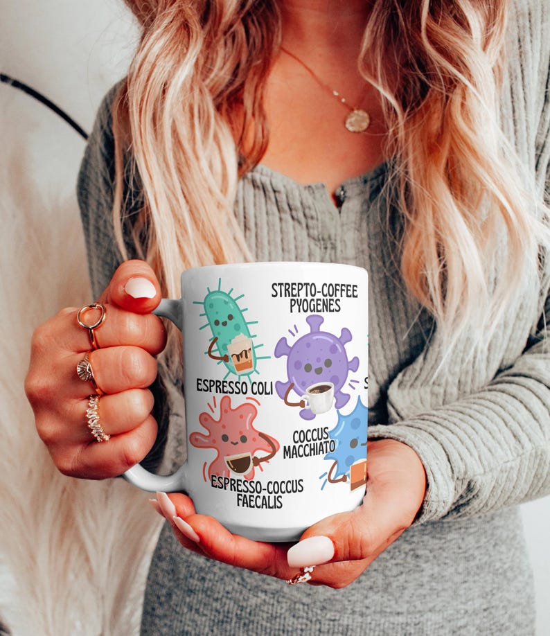 May include: A white ceramic coffee mug featuring cartoon illustrations of bacteria, each holding a coffee cup. The mug includes the text "Strepto-Coffee Pyogenes," "Espresso Coli," "Cocus Macchiato," and "Espresso-Cocus Faecalis."