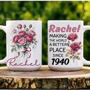 Puede incluir: Dos tazas de cerámica blanca con asas grandes. Una taza presenta un ramo de rosas rosas y el nombre "Rachel" en cursiva. La otra taza tiene el texto "Rachel MAKING THE WORLD A BETTER PLACE SINCE 1940" con acentos de rosas.