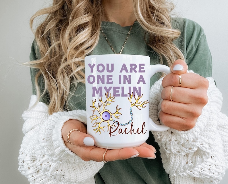May include: White ceramic mug featuring the text "YOU ARE ONE IN A MYELIN" in purple, a neuron illustration, and the name "Rachel" in cursive. The mug has a large handle. The mug is held by a person.