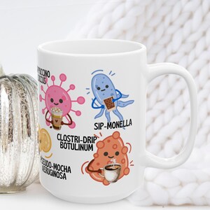 May include: White ceramic coffee mug featuring cartoon illustrations of microbes with coffee-themed puns. The mug includes the phrases "Cappuccino Coccus", "Sip-Monella", "Clostri-Drip Botulinum", and "Pseudo-Mocha Aeruginosa".