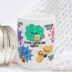 May include: White ceramic coffee mug featuring cartoon bacteria characters, each holding a cup of coffee. The mug has the words "Staphy-Cappuccino Aureus" and other coffee-themed puns. A decorative silver pumpkin is to the left.