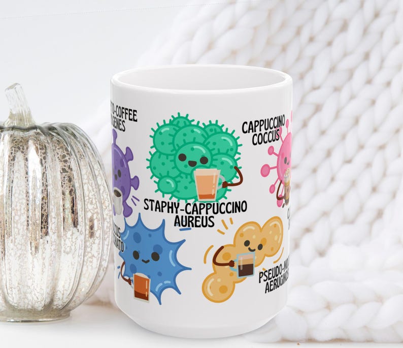 May include: White ceramic coffee mug featuring cartoon bacteria characters, each holding a cup of coffee. The mug has the words "Staphy-Cappuccino Aureus" and other coffee-themed puns. A decorative silver pumpkin is to the left.