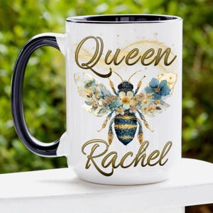 May include: White ceramic mug with a black handle and rim. The mug features a golden design with the words "Queen Rachel" and a bee illustration with floral wings in shades of blue, gold, and white.