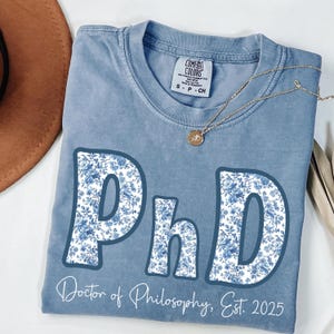 May include: A blue Comfort Colors t-shirt with the letters "PhD" in a floral pattern. The words "Doctor of Philosophy, Est. 2025" are printed below the letters. A gold necklace with a pendant is draped on the shirt.