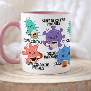 May include: White ceramic mug with a pink handle and rim, featuring cartoon illustrations of bacteria enjoying coffee. Text includes "Espresso Coli," "Strepto-Coffee Pyogenes," and "Espresso-Cocus Faecalis."