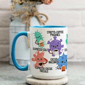 May include: White ceramic mug with a blue interior and handle. The mug features cartoon illustrations of bacteria, each holding a coffee cup or espresso. Text includes "Strepto-Coffee Pyogenes" and "Espresso Coli".