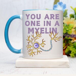 May include: White ceramic mug with a blue handle and rim. The mug features the text "YOU ARE ONE IN A MYELIN" in purple, along with a yellow and blue neuron illustration. The mug is sitting on a white surface.