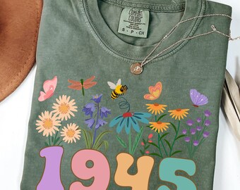 Può includere: T-shirt verde oliva con un design floreale con farfalle, un'ape e il numero "1945" in un carattere retrò colorato. La maglietta ha un'etichetta Comfort Colors ed è abbinata a una collana d'argento.