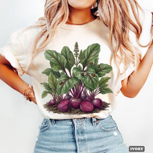 May include: Ivory-colored t-shirt featuring a detailed illustration of beets with large green leaves, purple roots, and ladybugs. The design includes sliced beets, showcasing the inner rings. The word "IVORY" is printed at the bottom right.