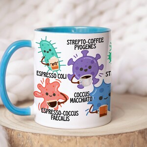 May include: White ceramic mug with a blue handle and interior. The mug features cartoon illustrations of bacteria, each holding a coffee cup, with text labels like "Espresso Coli" and "Strepto-Coffee Pyogenes".
