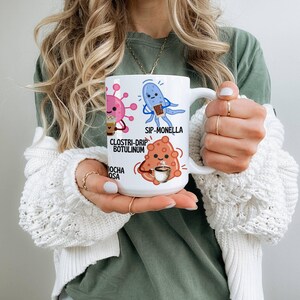 May include: White ceramic coffee mug featuring cartoon illustrations of microbes with coffee-related puns. The mug has the phrases "Clostri-drip Botulinum", "Sip-Monella", and "Mocha Osa" printed on it.