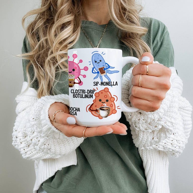 May include: White ceramic coffee mug featuring cartoon illustrations of microbes with coffee-related puns. The mug has the phrases "Clostri-drip Botulinum", "Sip-Monella", and "Mocha Osa" printed on it.