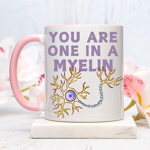 May include: White ceramic mug with a pink handle and interior. The mug features the text "YOU ARE ONE IN A MYELIN" in purple, along with a yellow and blue neuron illustration. The mug is on a white surface.