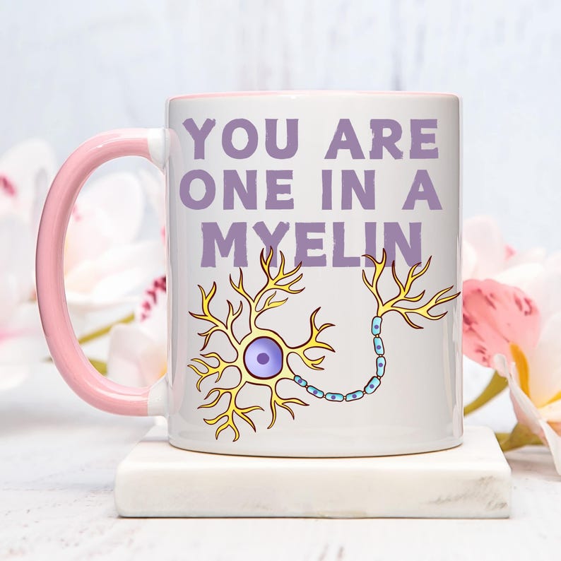 May include: White ceramic mug with a pink handle and interior. The mug features the text "YOU ARE ONE IN A MYELIN" in purple, along with a yellow and blue neuron illustration. The mug is on a white surface.