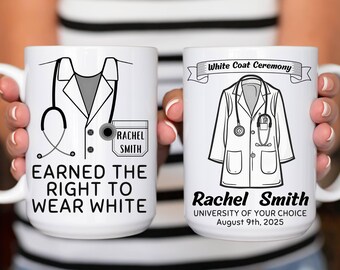 Könnte beinhalten: Zwei weiße Keramikbecher mit schwarz-weißen medizinischen Motiven. Ein Becher zeigt eine Laborkittel-Illustration und den Text "Earned the Right to Wear White" mit einem Namen. Der andere Becher zeigt eine Laborkittel-Illustration mit dem Text "White Coat Ceremony" sowie einen Namen, eine Universität und ein Datum.