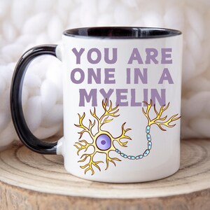 May include: White ceramic mug with a black interior and handle. The mug features the text "YOU ARE ONE IN A MYELIN" in purple, along with a yellow and blue neuron illustration. The mug is on a wooden surface.