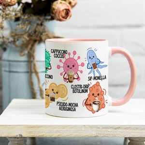 May include: White ceramic mug with a pink interior and handle, featuring cartoon illustrations of bacteria with coffee-themed names like "Cappuccino Coccus" and "Sip-Monella." The mug is on a white wooden stand.