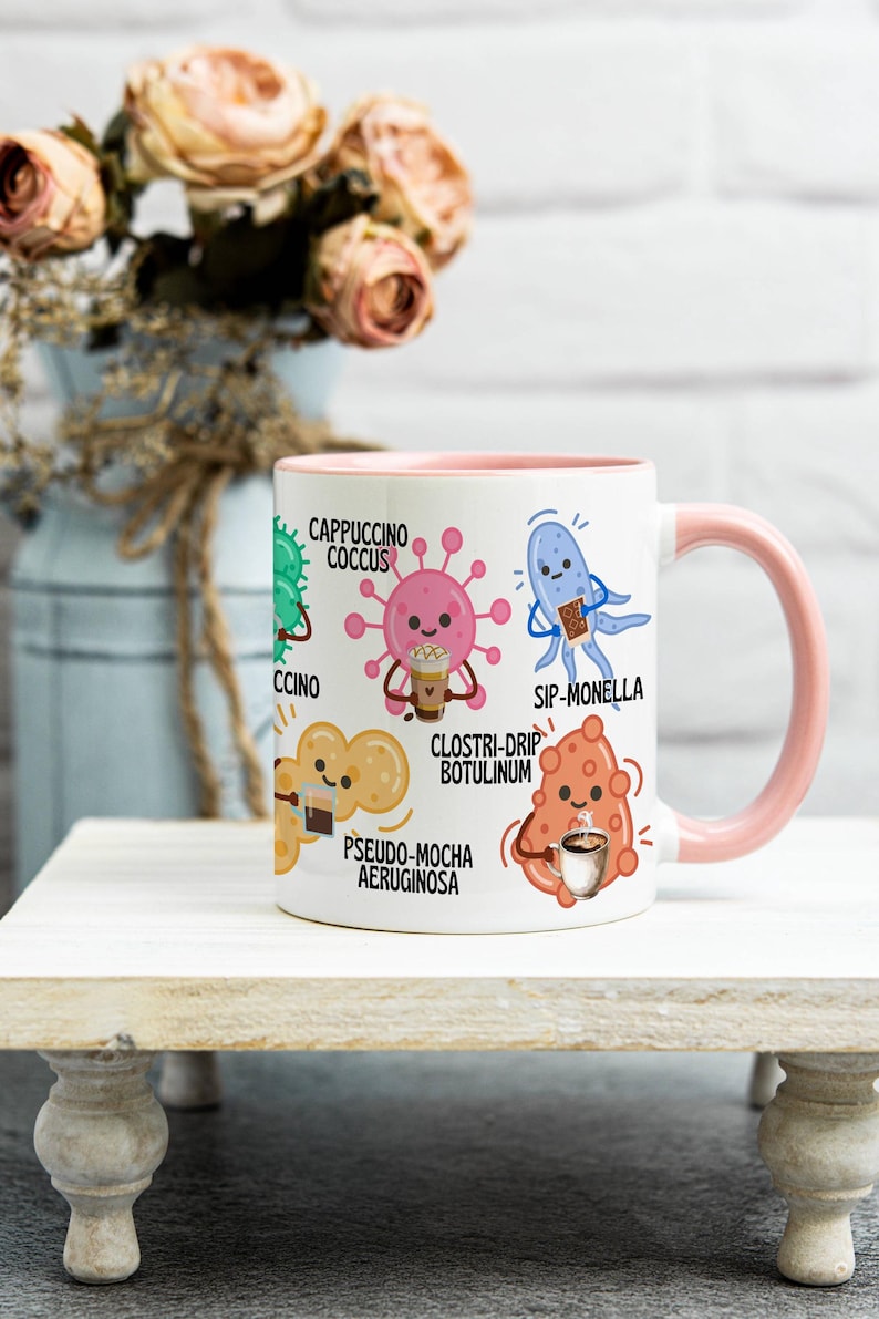 May include: White ceramic mug with a pink interior and handle, featuring cartoon illustrations of bacteria with coffee-themed names like "Cappuccino Coccus" and "Sip-Monella." The mug is on a white wooden stand.