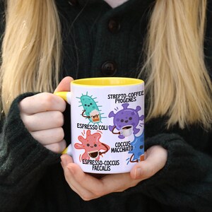 May include: A white ceramic mug with a yellow interior and handle, featuring cartoon illustrations of bacteria. The mug has the text "Espresso Coli", "Strepto-Coffee Pyogenes", "Coccus Macchiato", and "Espresso-Coccus Faecalis".
